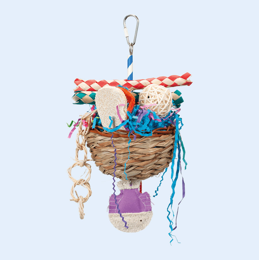 A hanging bird toy shaped like a basket, filled with colorful crinkle paper, woven bamboo tubes, loofah pieces, and a rattan ball. Designed for foraging and chewing, providing enrichment for parrots. Available at Squawk Shop.